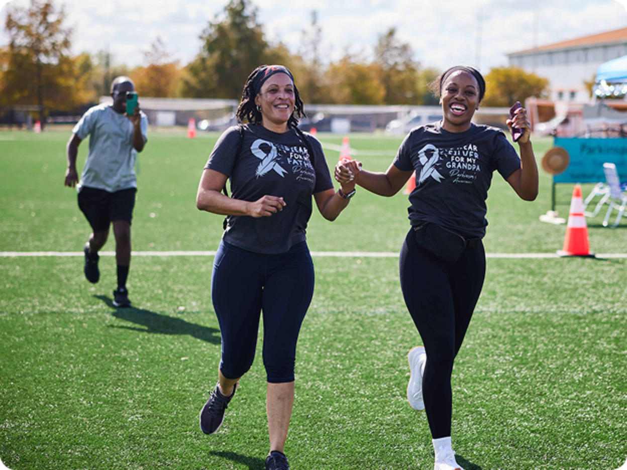 Two smiling women in matching memorial T-shirts run hand in hand across a grassy athletic field during an outdoor event.
