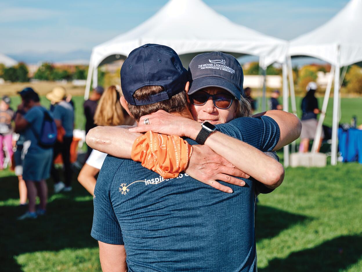 Two participants embrace at an outdoor Michael J. Fox Foundation event, with white tents and other attendees in the background.