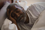 African American older man is asleep in bed. He wears a watch and is nestling his head with the other hand.