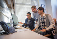 Three male researchers sitting or standing in front of a computer looking at a shared screen. 