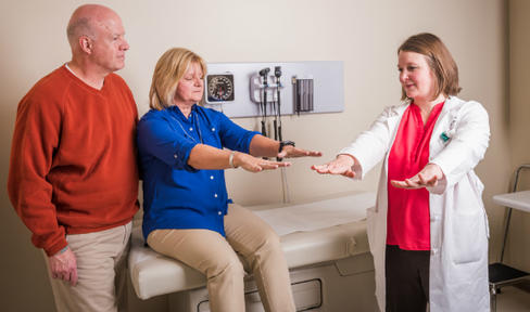 Male care partner alongside female patient undergoing Parkinson's test with her female physician.