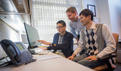 Three male researchers sitting or standing in front of a computer looking at a shared screen. 