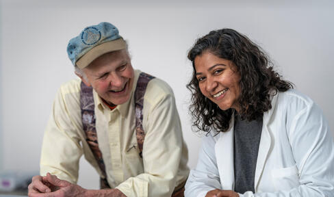 An older patient with a hat and yellow shirt laughs next to his smiling healthcare provider.