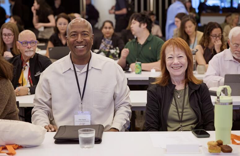 A man and woman smile in a crowded meeting room.