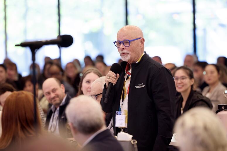 A man holds a microphone while standing in a crowded room.