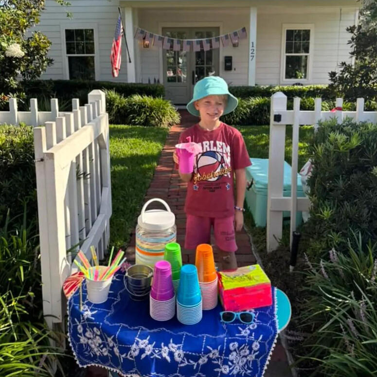 a young boy stands behind his lemonade stand, holding a cup