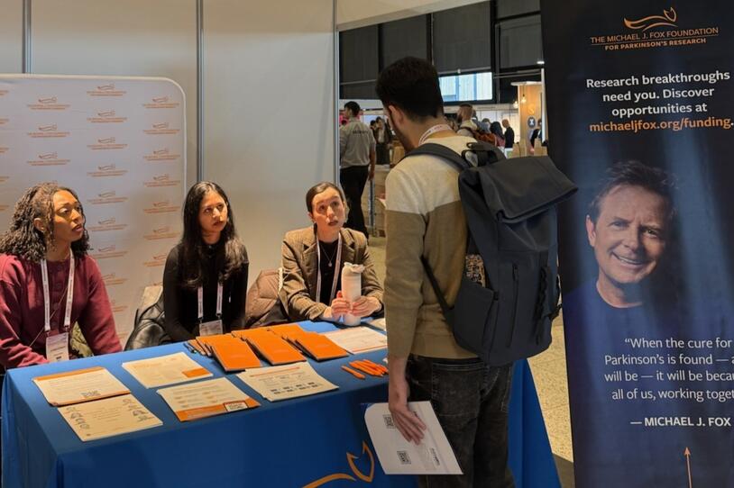 Three women sitting together behind a table with various pens, keychains, and informational papers on it, speaking with a man who is standing and facing the table. 