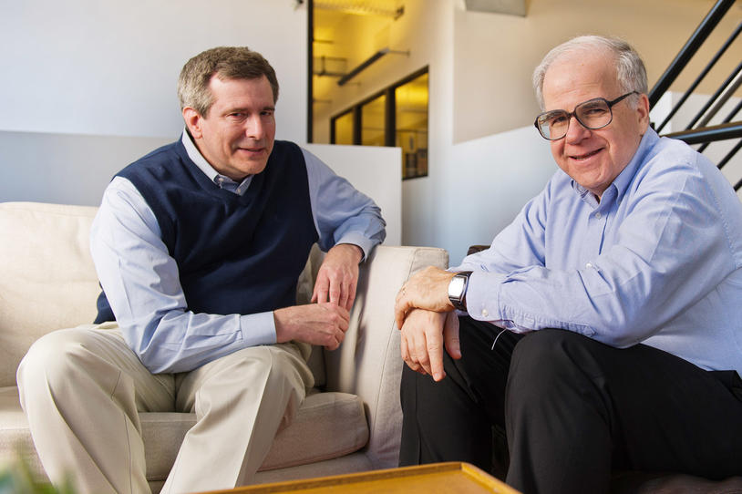 Two men sitting in a living area at work smiling for the camera.
