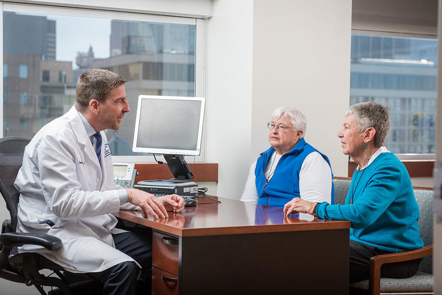 Male researcher sitting in at a desk talking with a female patient and care partner.