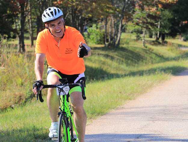 Male biker on country road smiling at camera with a thumbs up.