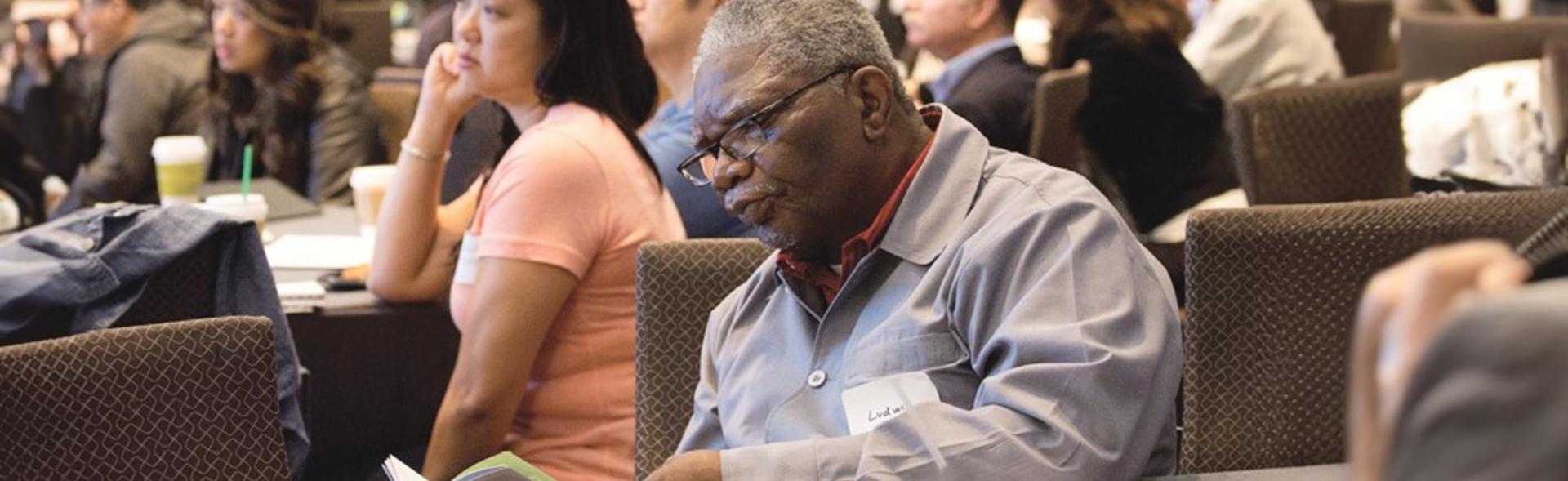 A man reading an informational pamphlet in a conference room with other people at an event