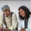 An older patient with a hat and yellow shirt laughs next to his smiling healthcare provider.
