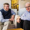 Two men sitting in a living area at work smiling for the camera.