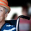 Older white woman wearing an orange striped fedora and blue and white striped shirt sitting in a wheelchair.