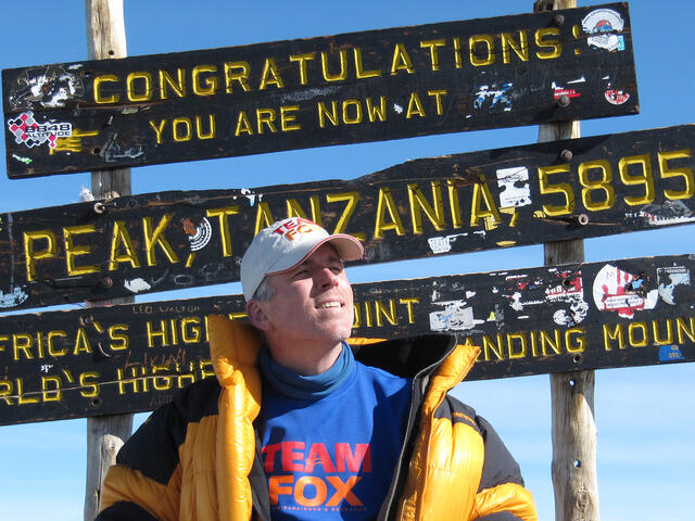 Tom Sabourin stands in front of the sign declaring the Uhuru peak of Mount Kilimanjaro, wearing a Team Fox shirt, and holding a book by Michael J. Fox.