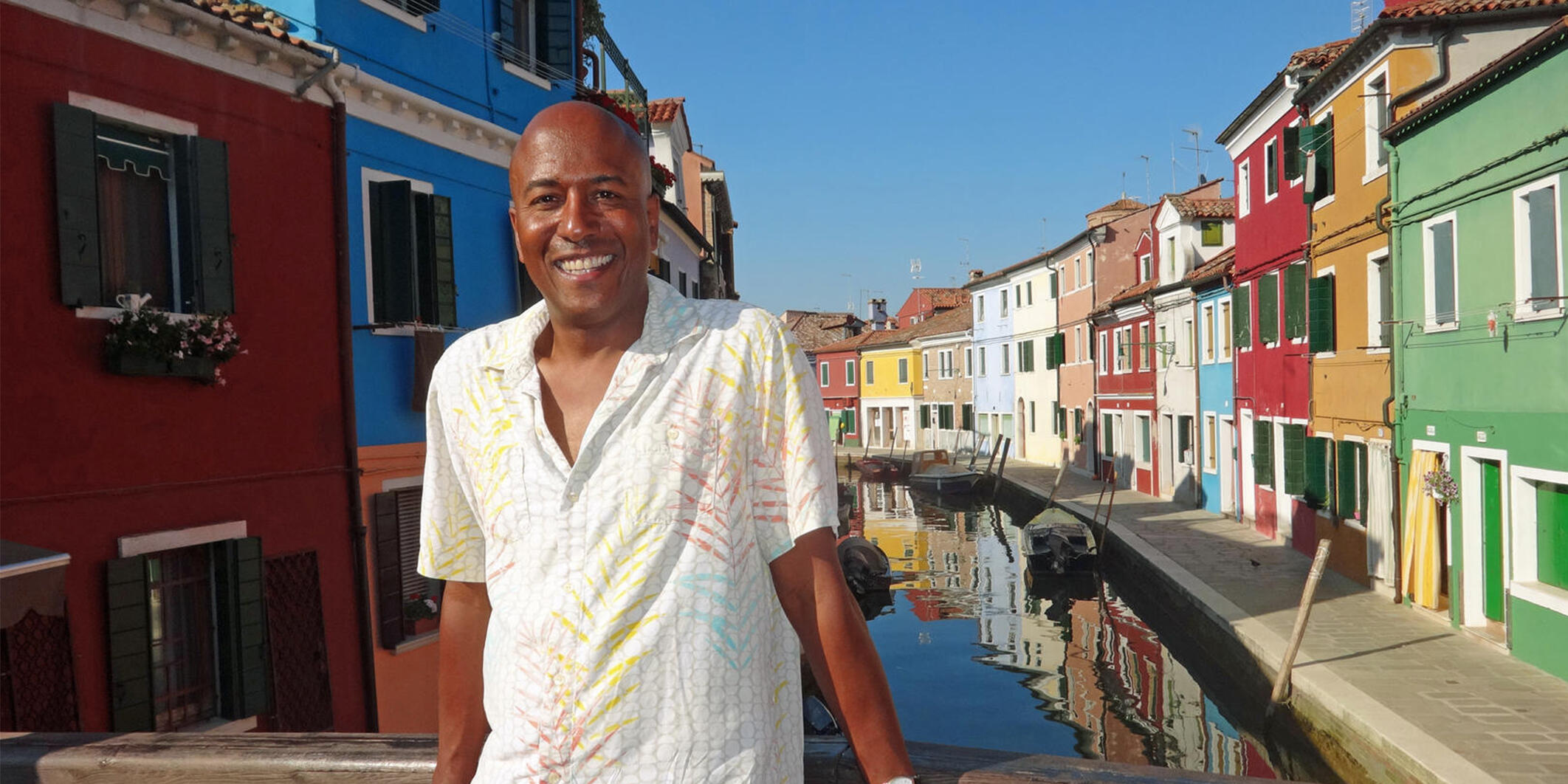 A man stands on a bridge over a canal lined with multi-colored buildings. 