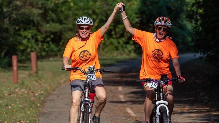 Two women riders in orange shirts raising joined hands as they pedal